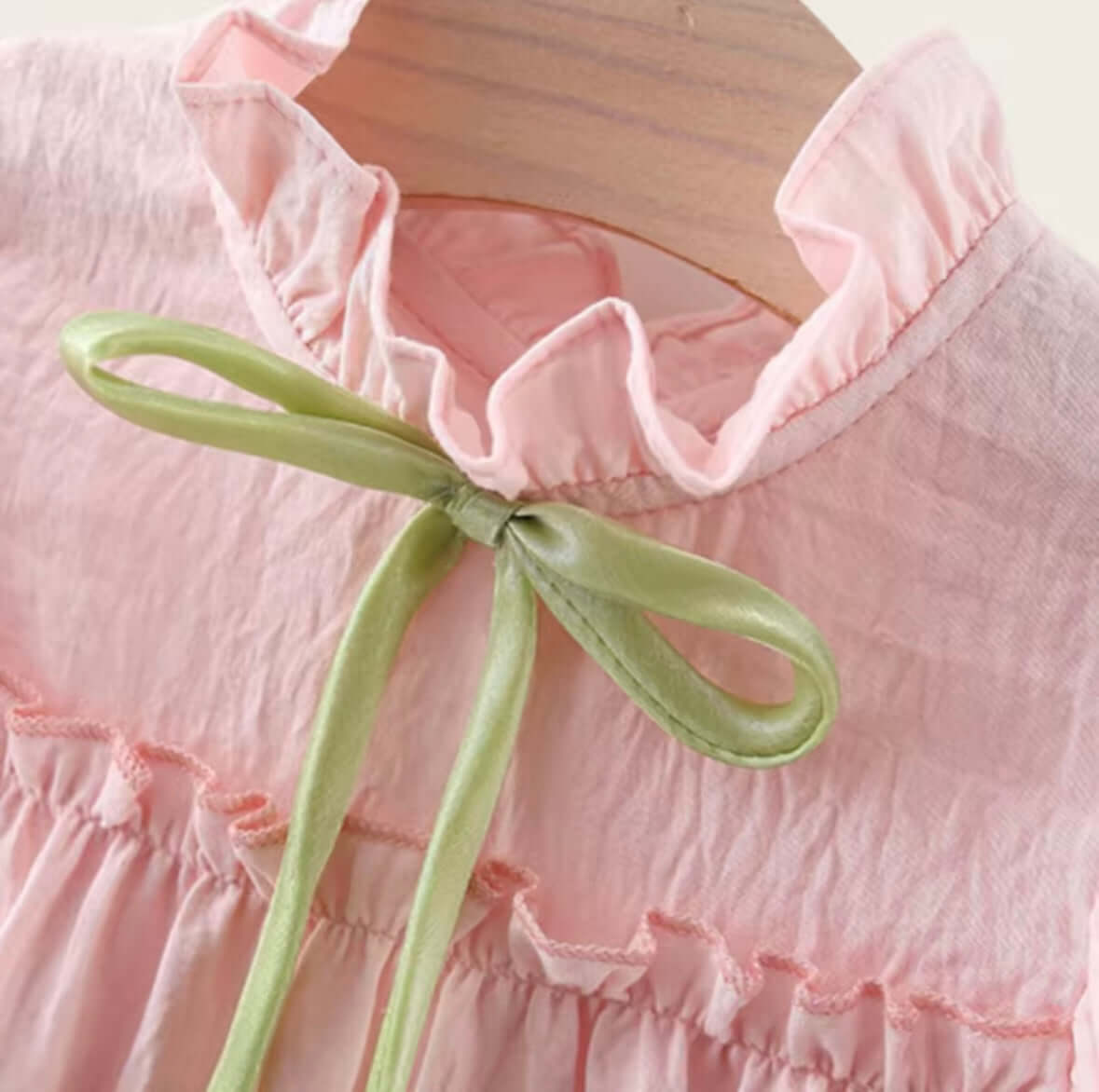 Close-up of blush pink ruffle dress with green bow tie and delicate tiered layers on wooden hanger.